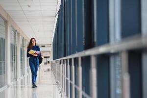 vrolijk brunette leerling meisje met zwart rugzak houdt boeken in modern gebouw. vrouw leerling staand met boeken in college gang foto