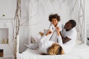 portret van gelukkig multiraciaal jong familie aan het liegen Aan knus wit bed Bij huis, glimlachen Internationale mam en vader ontspannende met weinig biraciaal meisje kind poseren voor afbeelding in slaapkamer foto