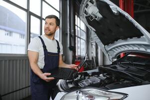 monteur Mens monteur manager arbeider gebruik makend van een laptop computer controle auto in werkplaats Bij auto auto reparatie onderhoud centrum. ingenieur jong Mens op zoek Bij inspectie voertuig details onder auto kap foto