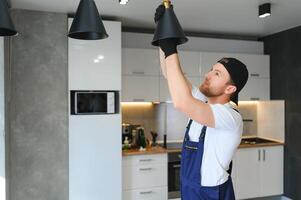 mannetje elektricien veranderingen de licht bollen in de lamp. een jong Mens in overall presteert technisch werk Aan elektrificatie van terrein. foto