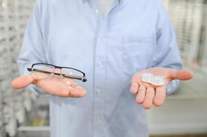 oogheelkunde concept. portret van Mens kiezen tussen bril en contact lenzen staand in de optisch op te slaan, Holding plastic geval en specificaties in beide handen, op zoek Bij eyewear foto