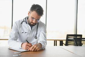 dokter Mens zittend Bij de bureau Bij zijn werken plaats. perfect medisch onderhoud in kliniek. portret van een knap jong dokter. foto