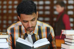 portret van vrolijk mannetje Internationale Indisch leerling met rugzak, aan het leren accessoires staand in de buurt boekenkasten Bij Universiteit bibliotheek of boek op te slaan gedurende breken tussen lessen. onderwijs concept foto