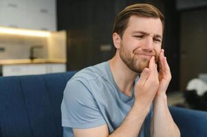 kiespijn. ongelukkig van streek Kaukasisch verdrietig Mens zit Aan de bankstel Bij huis, houdt zijn hand- in de buurt zijn wang, grimassen, heeft een acuut kiespijn, behoeften een overleg met een tandarts dokter foto