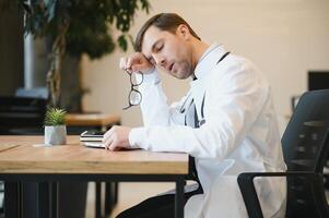 benadrukt mannetje dokter za Bij zijn bureau. midden volwassen mannetje dokter werken lang uur. overwerkt dokter in zijn kantoor. niet zelfs artsen zijn vrijgesteld van burn-out . foto