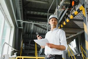 een jong geslaagd ingenieur met een tekening in zijn handen is staand in de gebied van een modern fabriek. foto