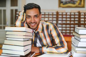 portret van vrolijk mannetje Internationale Indisch leerling met rugzak, aan het leren accessoires staand in de buurt boekenkasten Bij Universiteit bibliotheek of boek op te slaan gedurende breken tussen lessen. onderwijs concept foto