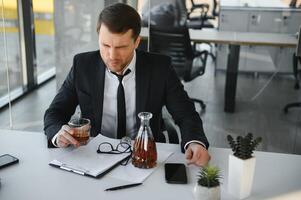 vermoeidheid jong zakenman zittend Bij werkplaats en Holding whisky fles, drinken alcohol, faillissement concept. foto