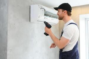 glimlachen jong mannetje elektricien reparatie conditioner in cliënt huis. Mens monteur of klusjesman fix filters in lucht staat apparaat Bij klant huis foto