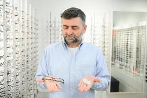 oogheelkunde concept. portret van Mens kiezen tussen bril en contact lenzen staand in de optisch op te slaan, Holding plastic geval en specificaties in beide handen, op zoek Bij eyewear foto