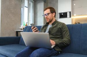 portret van een aantrekkelijk echt jong gebaard Mens vervelend gewoontjes kleren zittend Aan een bankstel Bij de leven kamer, pratend Aan mobiel telefoon terwijl gebruik makend van laptop computer. foto