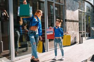 mooi mam en haar schattig weinig dochter zijn Holding boodschappen doen Tassen, op zoek Bij camera en glimlachen terwijl staand buitenshuis. boodschappen doen concept foto