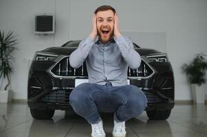 emotioneel knap vent verhogen hand- omhoog en glimlachen Bij camera. rijk Mens buying auto Bij luxe handel salon foto