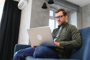 mannen werken Aan laptop computer in zijn kamer. huis werk of studie, freelance concept. jong Mens zittend ontspannen Aan sofa met laptop. modern zakenman gebruik makend van laptop. foto