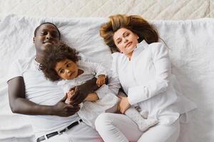 portret van gelukkig multiraciaal jong familie aan het liegen Aan knus wit bed Bij huis, glimlachen Internationale mam en vader ontspannende met weinig biraciaal meisje kind poseren voor afbeelding in slaapkamer foto