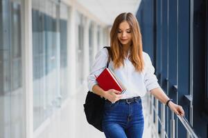 vrolijk brunette leerling meisje met zwart rugzak houdt boeken in modern gebouw. vrouw leerling staand met boeken in college gang foto