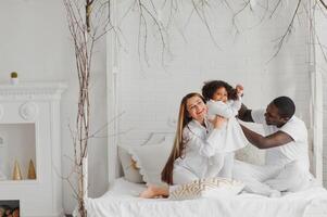 portret van gelukkig multiraciaal jong familie aan het liegen Aan knus wit bed Bij huis, glimlachen Internationale mam en vader ontspannende met weinig biraciaal meisje kind poseren voor afbeelding in slaapkamer foto