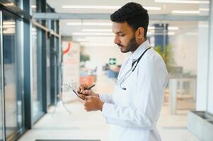 portret van gelukkig vriendelijk mannetje Indisch Latijns dokter medisch arbeider vervelend wit jas met stethoscoop in de omgeving van nek staand in modern privaat kliniek op zoek Bij camera. medisch gezondheidszorg concept foto