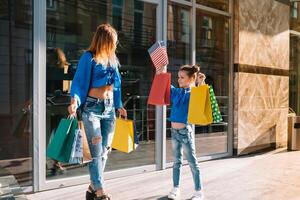 mooi mam en haar schattig weinig dochter zijn Holding boodschappen doen Tassen, op zoek Bij camera en glimlachen terwijl staand buitenshuis. boodschappen doen concept foto