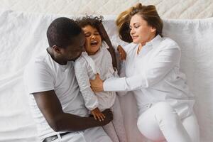 portret van gelukkig multiraciaal jong familie aan het liegen Aan knus wit bed Bij huis, glimlachen Internationale mam en vader ontspannende met weinig biraciaal meisje kind poseren voor afbeelding in slaapkamer foto