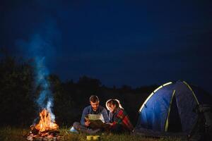 camping nacht in bergen. toerist paar zittend in voorkant van verlichte tent lit door brandend kampvuur. toerisme en buitenshuis werkzaamheid concept. foto