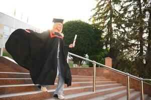 gelukkig schattig Kaukasisch grad meisje is lachend. ze is in een zwart Mortier bord, met rood kwast, in gewaad, met mooi hoor bruin gekruld haar, diploma in hand- foto