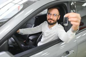 knap jong zakenman in klassiek wit pak is lachend, op zoek Bij camera en tonen auto sleutels terwijl zittend in auto in een motor show. foto