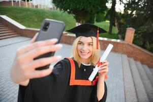gelukkig vrouw leerling Holding Universiteit diploma terwijl nemen selfie na diploma uitreiking ceremonie. foto