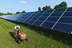 een Mens werken Bij zonne- macht station. een arbeider Aan een tuin trekker maait gras Aan een zonne- paneel boerderij. foto
