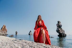 vrouw reizen zee. gelukkig toerist in rood jurk genieten nemen afbeelding buitenshuis voor herinneringen. vrouw reiziger poseren Aan de rots Bij zee baai omringd door vulkanisch bergen, sharing reizen avontuur reis foto