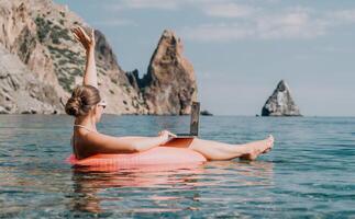 vrouw freelancer werken Aan laptop zwemmen in zee Aan roze opblaasbaar ring. gelukkig toerist in zonnebril drijvend Aan opblaasbaar donut en werken Aan laptop computer in kalmte oceaan. afgelegen werken overal foto