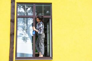 een vrouw handmatig wast de venster van de huis met een vod met een verstuiven schoonmaakster en een dweilen buiten. veiligheid Bij hoogte, herstellen bestellen en netheid in de lente, schoonmaak onderhoud foto