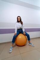 jong meisje zittend Aan oranje geschiktheid bal. vrouw in jeans leert oefeningen met een bal. foto