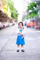 portret van aziatisch leerlingenmeisje dat blauw en wit schooluniform draagt. kind meisje schaamt zich een beetje als je ouders een foto maken als de baby voor het eerst naar school gaat. lief lachend kind van 3 jaar oud.