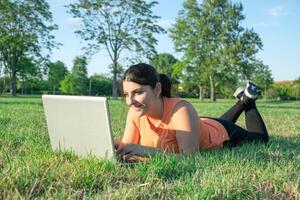 jong Kaukasisch vrouw aan het liegen Aan park gebruik makend van haar laptop heel gelukkig foto