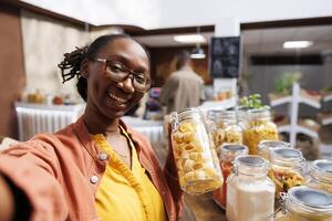 in bio-voedsel winkel, zwart vrouw vervelend bril houdt glas houder geladen met spaghetti. biologisch producten, vloggen, digitaal camera, en klant videografie zijn allemaal voorbeelden van online marketing. foto