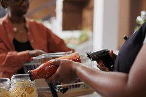 gedetailleerd schot presentatie van een Afrikaanse Amerikaans Kassa scannen een fles van eigengemaakt pasta saus voor klanten. een dichtbij visie van een vrouw verkoper gebruik maken van een streepjescode scanner Bij de uitchecken balie. foto