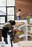 in bio voedsel markt, vrouw verkoper vervelend een schort organiseert van eigen bodem fruit en groenten Aan planken. in eco vriendelijk kruidenier op te slaan, Afrikaanse Amerikaans verkoper inspecteert vers biologisch produceren. foto