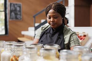 portret van zwart vrouw vervelend een schort werken in eco vriendelijk kruidenier op te slaan, regelen van biologisch duurzame artikelen. Afrikaanse Amerikaans verkoper organiserende glas potten gevulde met gezond bewaard gebleven artikelen. foto