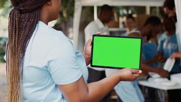 jong zwart vrouw geklemd digitaal tablet met geïsoleerd mockup sjabloon voor liefdadigheid berichten. Afrikaanse Amerikaans vrijwilliger draag- slim apparaatje met blanco groen scherm. over- de schouder statief. foto