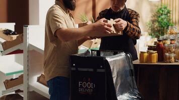 postbode het verstrekken van biologisch nul verspilling supermarkt voedsel bestellingen naar klanten, hielpen door ouderen kleinhandel klerk naar vullen warmte- rugzak. Mens brengen eco vriendelijk lokaal winkel boodschappen naar klanten foto