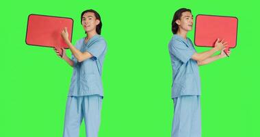 medisch verpleegster presenteert toespraak bubbel in studio met groene scherm achtergrond, Holding karton icoon met geïsoleerd kopieerruimte. gezondheidszorg arbeider tonen klein aanplakbord teken Aan camera. foto