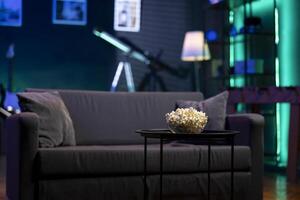 kom van popcorn Aan koffie tafel in voorkant van knus meubilair in leeg neon lit modern appartement. tussendoortje bereid voor film nacht in vaag verlichte huis theater in voorkant van bankstel foto