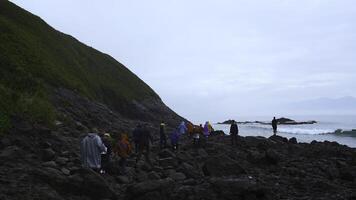 groep van toeristen wandelingen Aan steen oever. klem. toeristen wandelen Aan gevaarlijk steen kust in bewolkt het weer. groep van toeristen Aan kust met zwart rotsen van noordelijk landschap foto