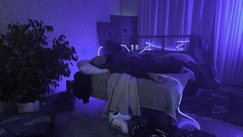 jong Mens gebruik makend van smartphone aan het liegen Aan bed Bij slaapkamer. media. knus bachelor opleiding kamer interieur met neon verlichting. foto