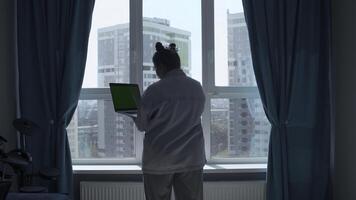 achterzijde visie van vrouw in pyjama Bij huis staand door de venster en typen. media. jong vrouw werken Aan laptop met groen scherm. foto