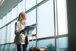 gelukkig jong vrouw Aziatisch is staan in luchthaven in de buurt koffer en lezing kaart foto