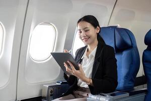 glimlachen gelukkig bedrijf vrouw Aziatisch vliegend en werken in een vliegtuig in eerste klas, vrouw zittend binnen een vliegtuig gebruik makend van digitaal tablet foto