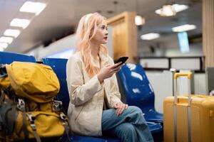 gelukkig Aziatisch toerist vrouw gebruik makend van mobiel smartphone met koffer op reis tussen wacht voor vlucht in luchthaven terminal, vlucht controleren in, toerist reis reis concept foto