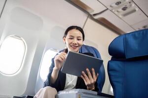 glimlachen gelukkig bedrijf vrouw Aziatisch vliegend en werken in een vliegtuig in eerste klas, vrouw zittend binnen een vliegtuig gebruik makend van digitaal tablet foto